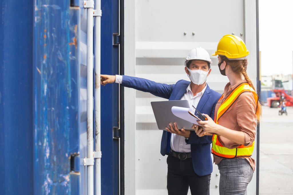 Teamwork foreman man and woman wearing protection face mask and safety helmet using laptop and holding clipboard checking containers in cargo ship for import export, Industrial container cargo concept.